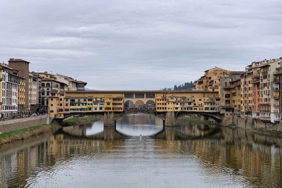 A historic bridge with shops spans a calm river, with reflections of the buildings and an overcast sky above.