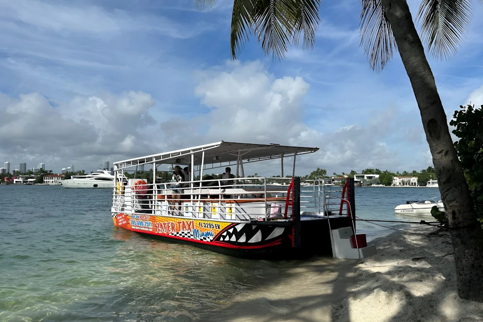 A colorful water taxi, with passengers on board, docked on a sandy shore under a sunny, partly cloudy sky, with palm trees overhead.