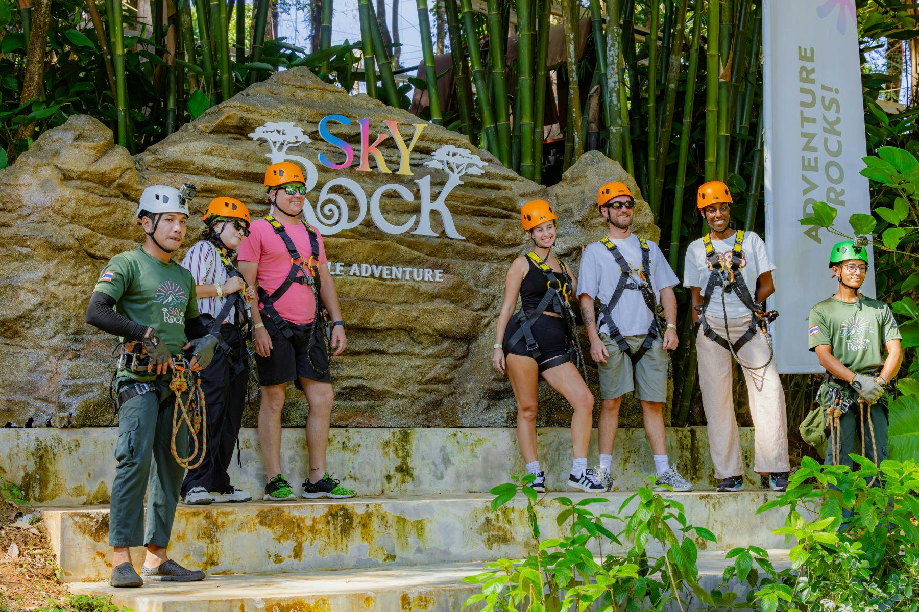 A group of people wearing helmets and harnesses stand on stone steps in front of a sign that says "Sky Rock Jungle Adventure."