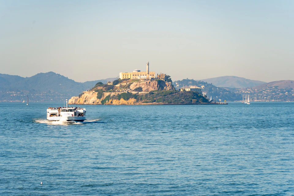 A distant view of Alcatraz Island with its iconic buildings, surrounded by water, and a ferry sailing nearby.