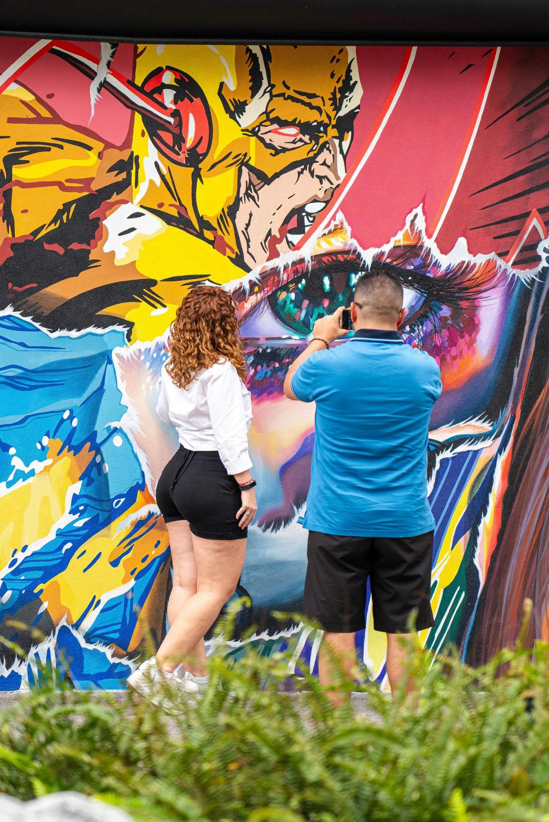 Two people stand in front of a colorful graffiti mural; one is taking a photo with a smartphone.