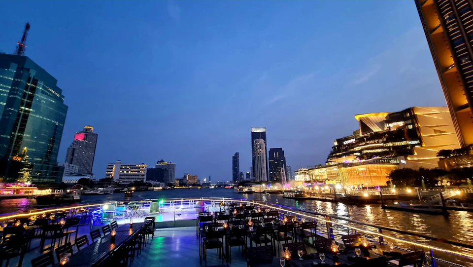 Outdoor restaurant on a boat with views of a city skyline and river. Tables are set with lit candles, and buildings are illuminated.
