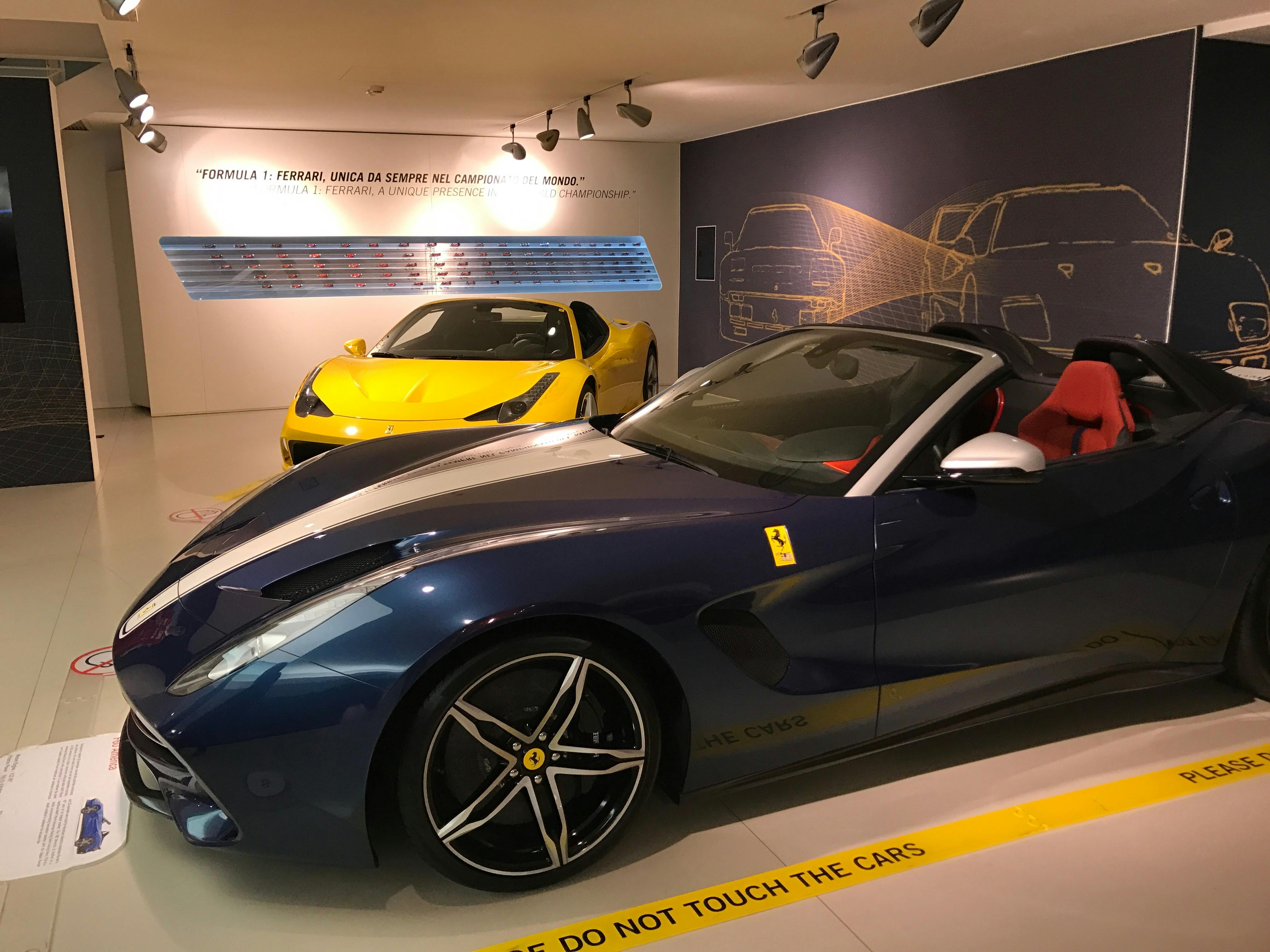 노란색 페라리와 파란색 페라리 컨버터블이 벽에 자동차 그래픽이 있는 쇼룸에 전시되어 있습니다.