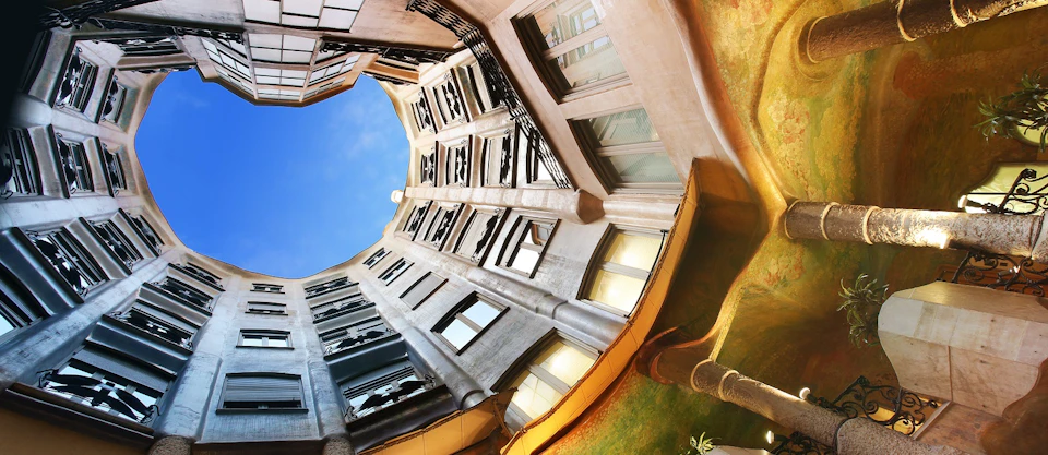 Looking up from the courtyard of a building, showing surrounding windows, decorative columns, and a clear blue sky through the opening.