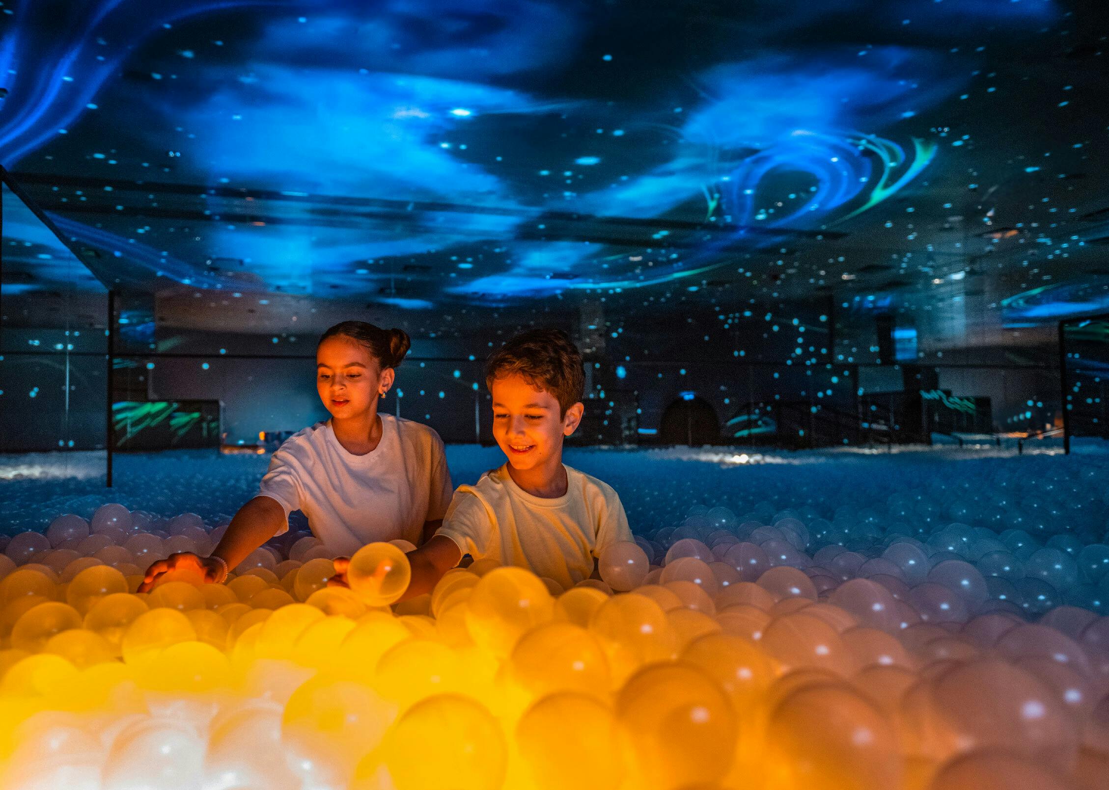 Two children in a glowing ball pit with a colorful ceiling display resembling a starry sky.
