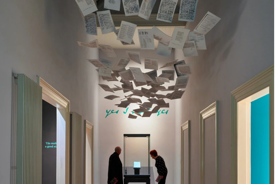 Two people observe a display in a hallway with suspended sheets of paper and framed art on side walls.