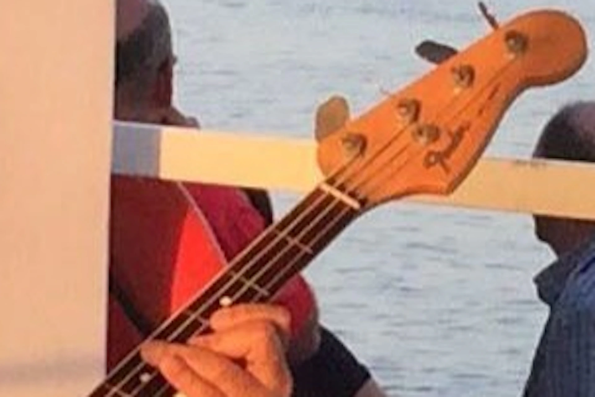 Three musicians play instruments—bass guitar, trumpet, and saxophone—on a boat with a river and distant shoreline in the background.