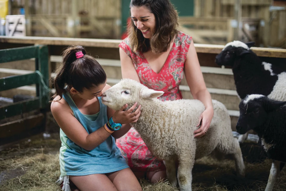 A girl and a woman smiling as the girl gently holds a lamb in a barn setting.