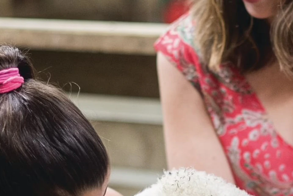 A girl and a woman smiling as the girl gently holds a lamb in a barn setting.