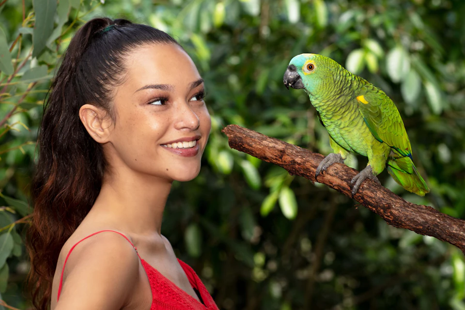 A woman in a red dress smiles at a green parrot perched on a branch, with a leafy background.