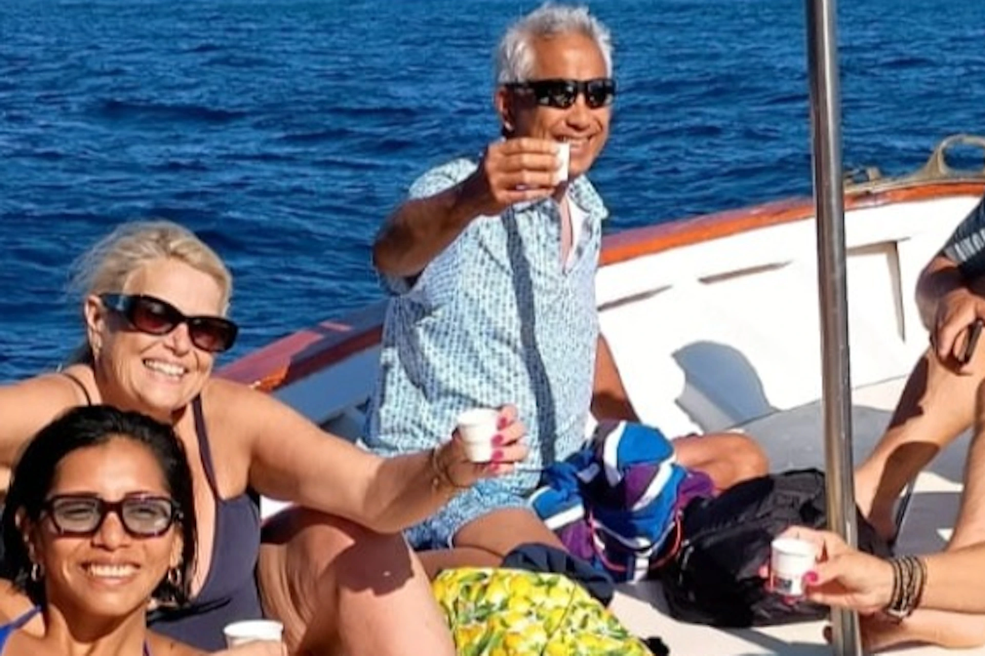 A group of people on a boat at sea, smiling and toasting with drinks, with an Italian flag flying in the background.