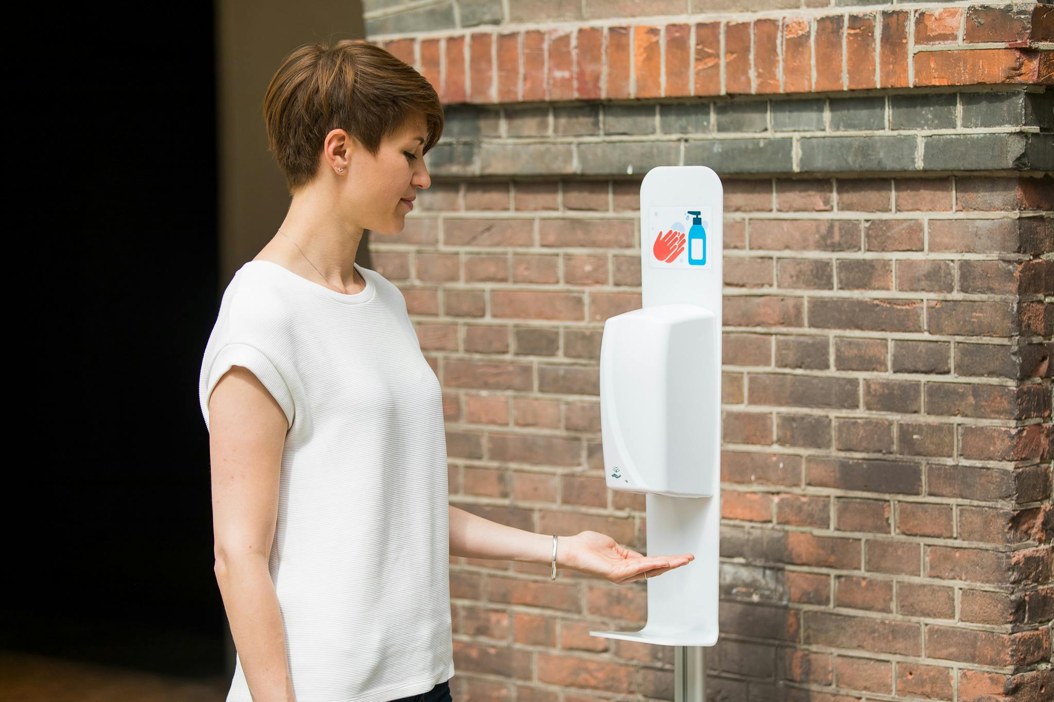 A person in a white shirt uses a wall-mounted hand sanitizer dispenser against a brick wall.