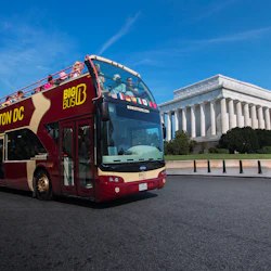 A double-decker tour bus drives near the Lincoln Memorial under a clear blue sky.