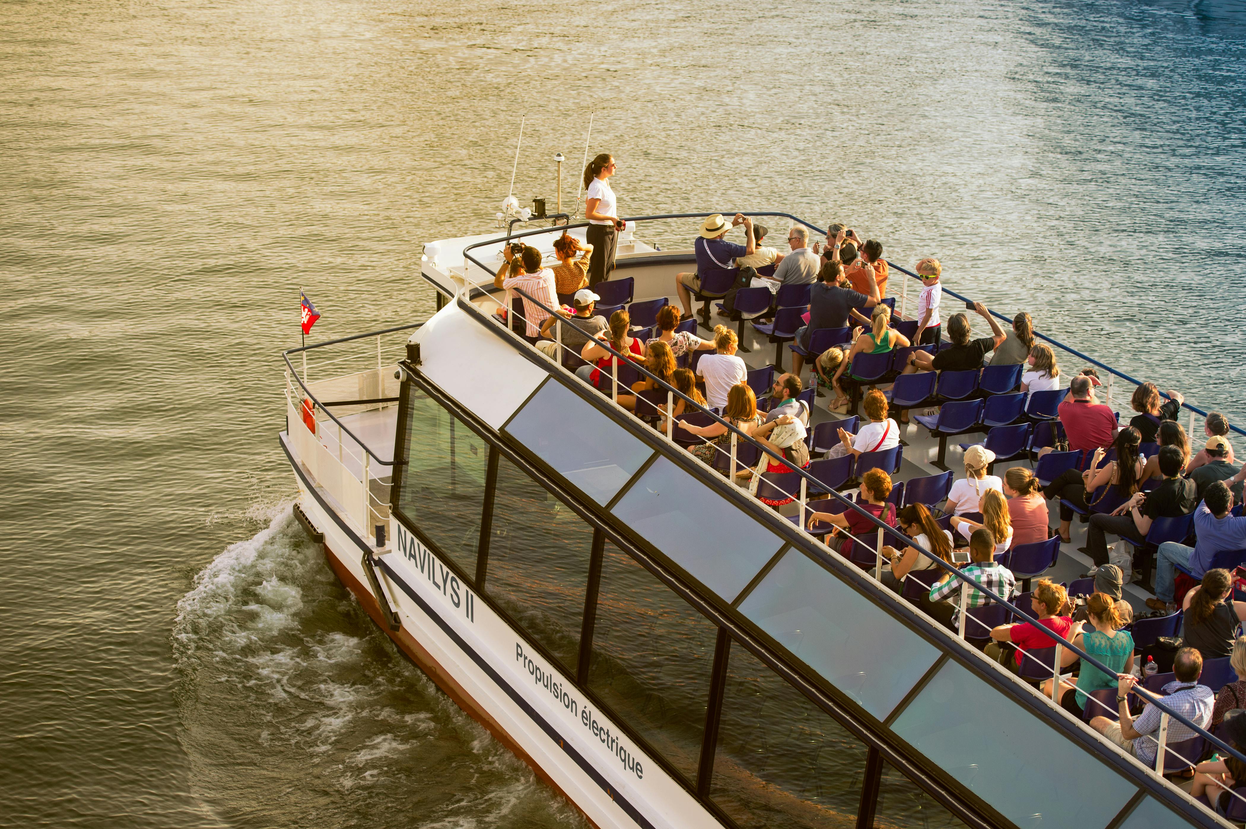 A boat filled with people in blue seats sailing on water; a woman stands near the helm, and passengers take photos.