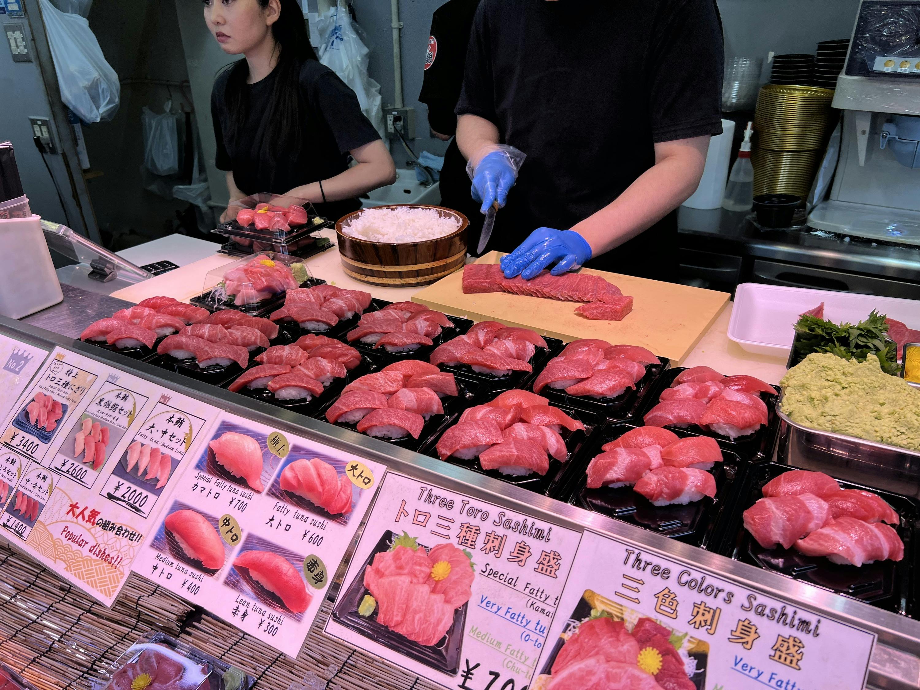 A sushi stall with assorted sashimi on display, staff preparing fish behind the counter.