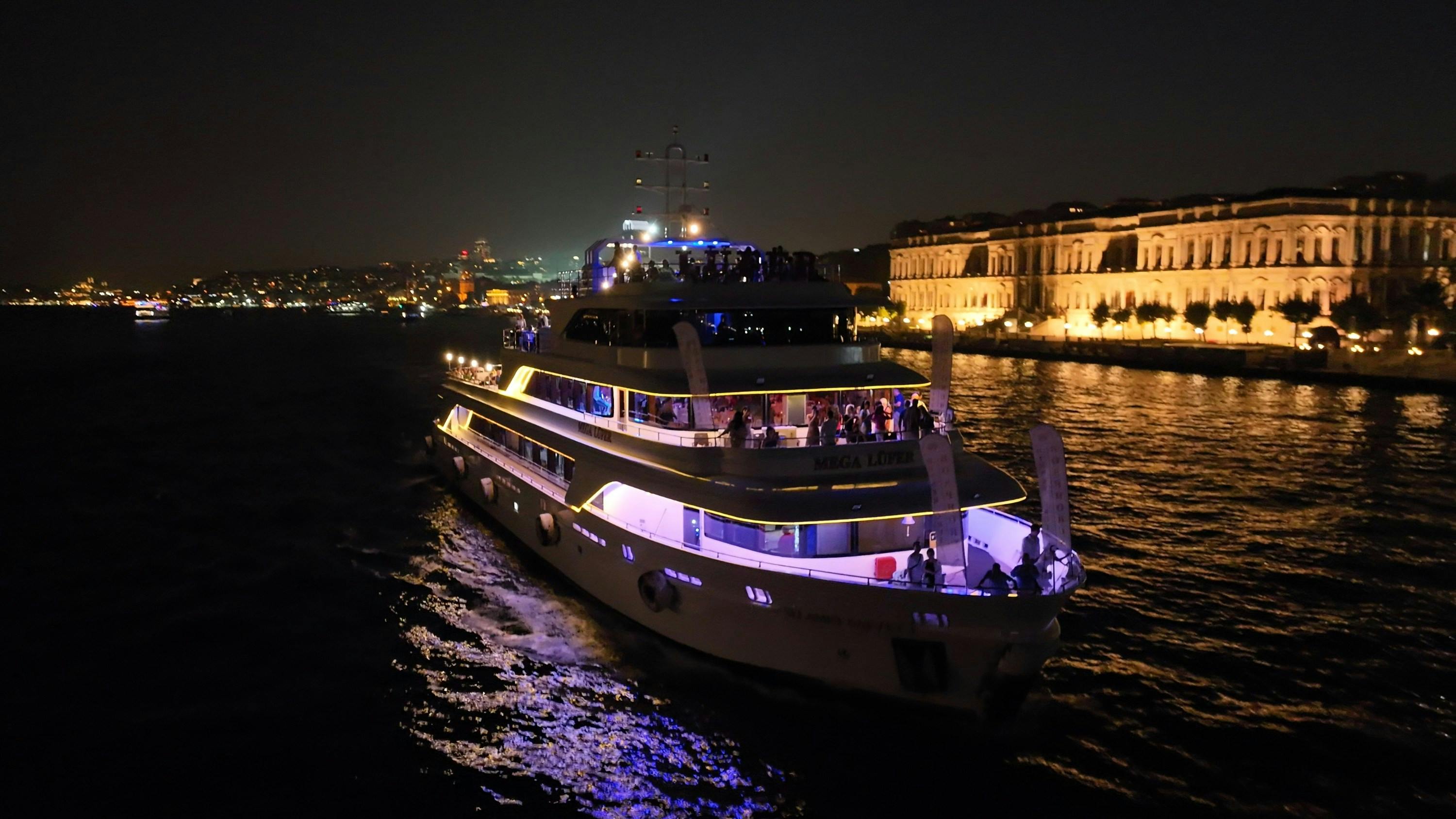 A large yacht illuminated with lights, cruising at night on water, with a lit cityscape and buildings in the background.