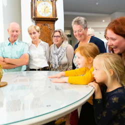 Grupp av vuxna och barn som observerar en utställning på ett glasbord i ett museum, med en fågelbur och klocka i bakgrunden.