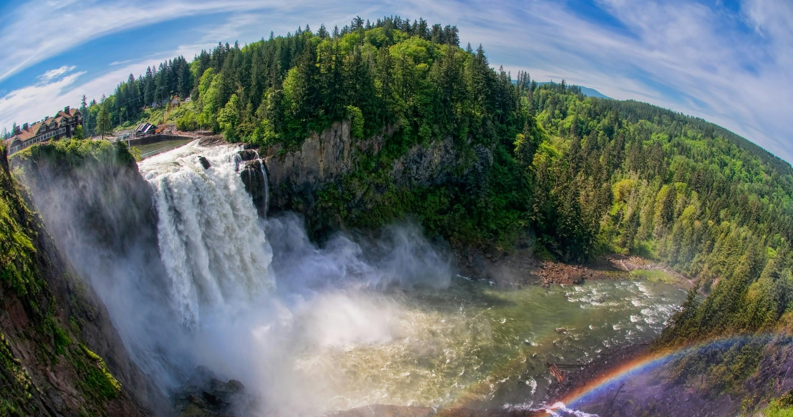 Ein Wasserfall, der über eine Klippe stürzt, umgeben von dichtem grünen Wald, mit Nebel und einem schwachen Regenbogen.