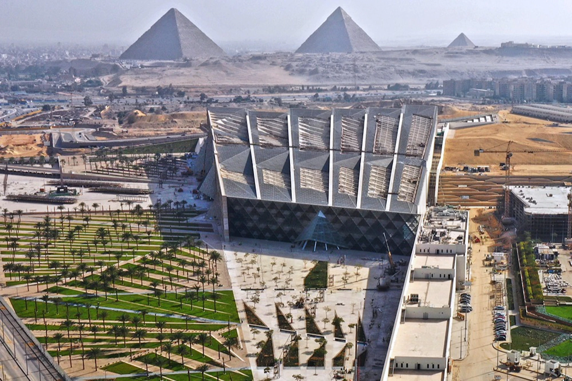A modern, angular building with extensive gardens in front, set against a backdrop of three pyramids in the distance.