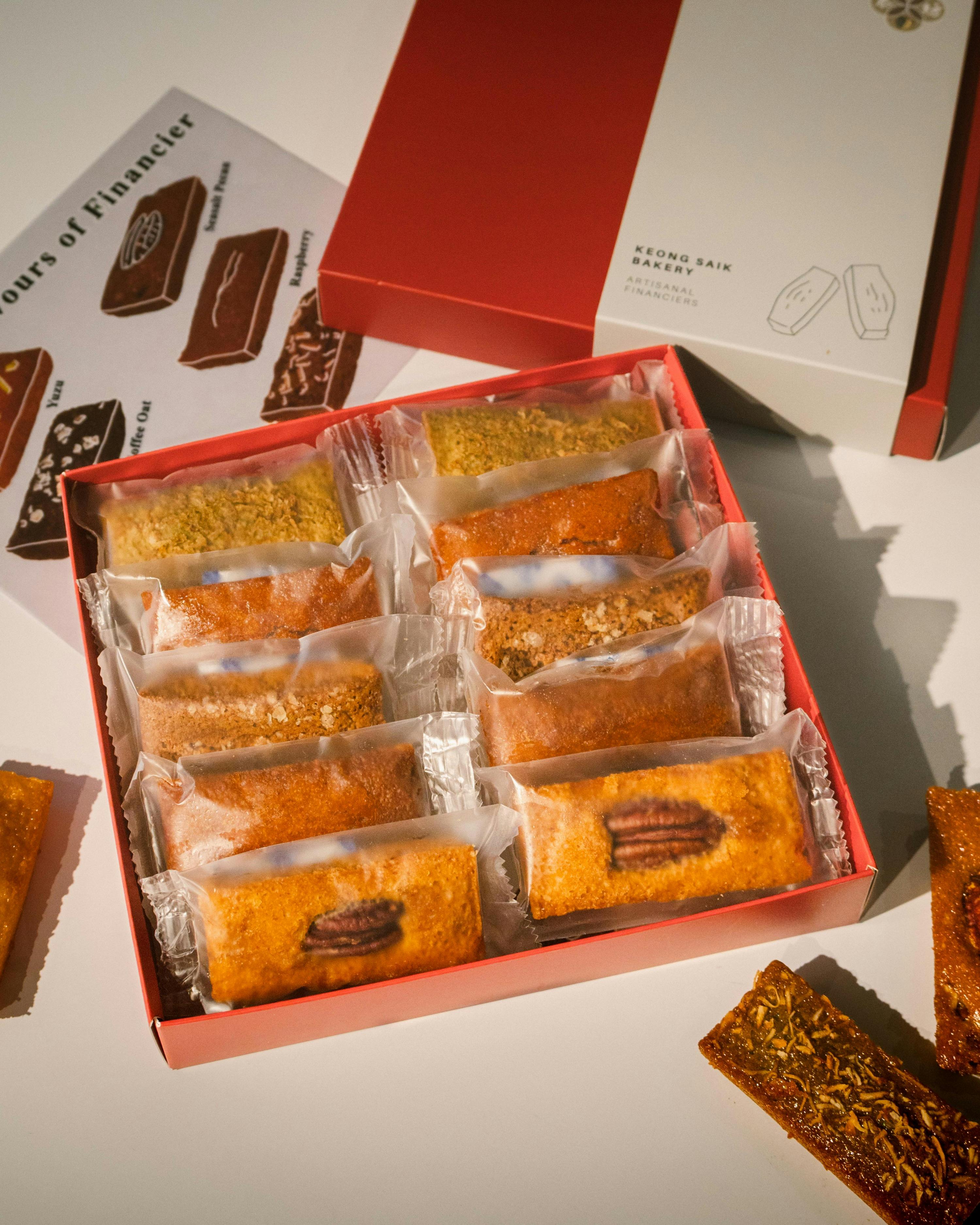 A box of assorted packaged financiers displayed on a table, with a red box and decorative paper in the background.