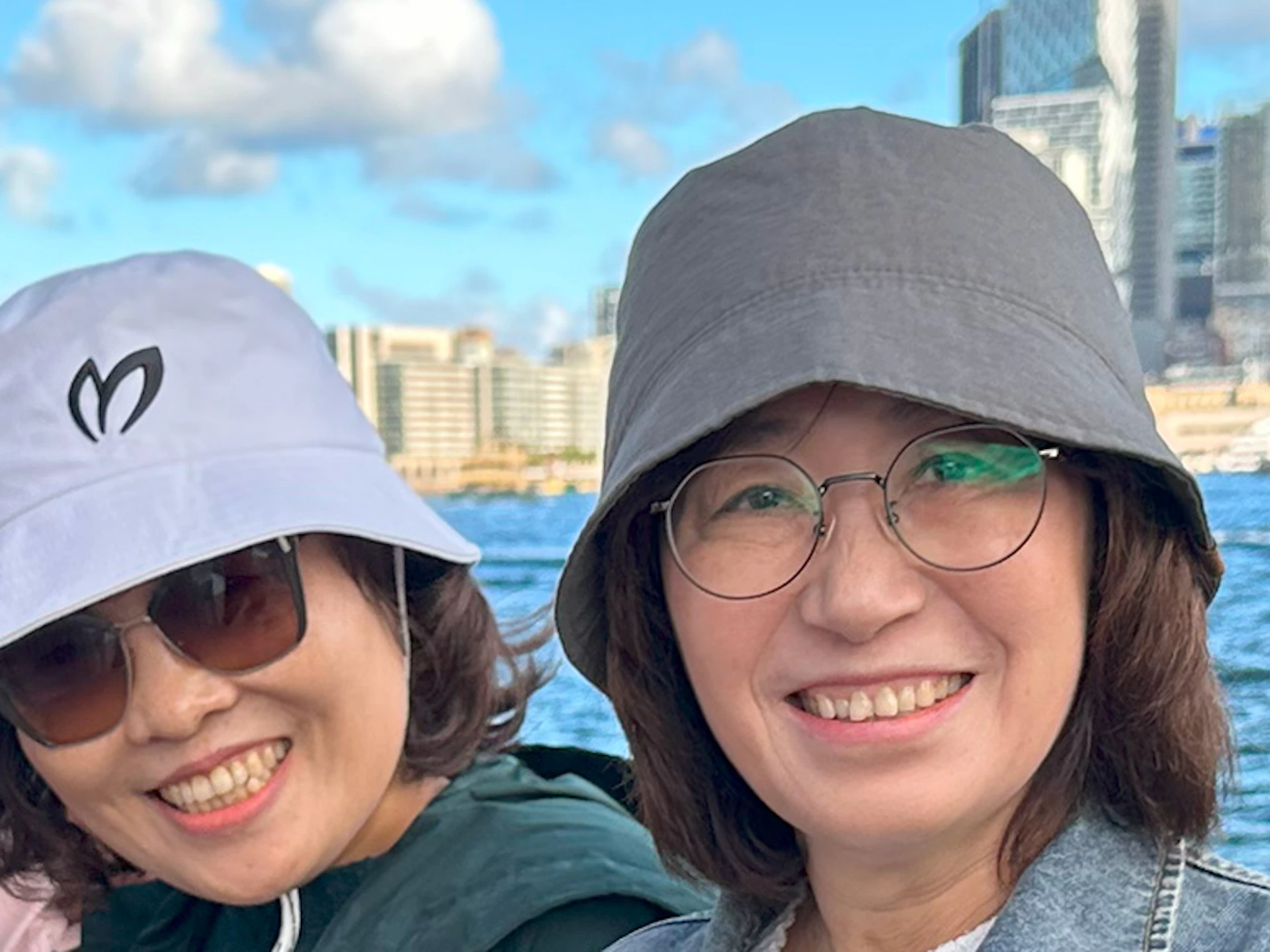 A group of smiling people posing outdoors with a city skyline, a bridge, and water in the background on a sunny day.