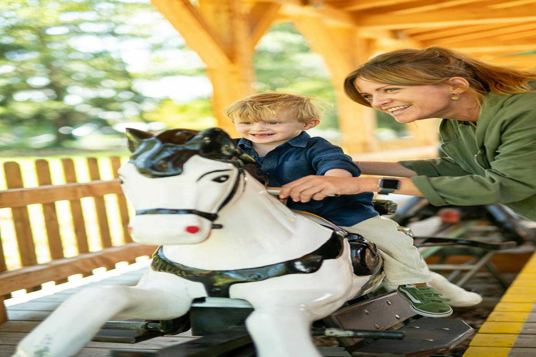 A child rides a white carousel horse while a smiling adult assists, under a wooden structure with sunlight filtering through.