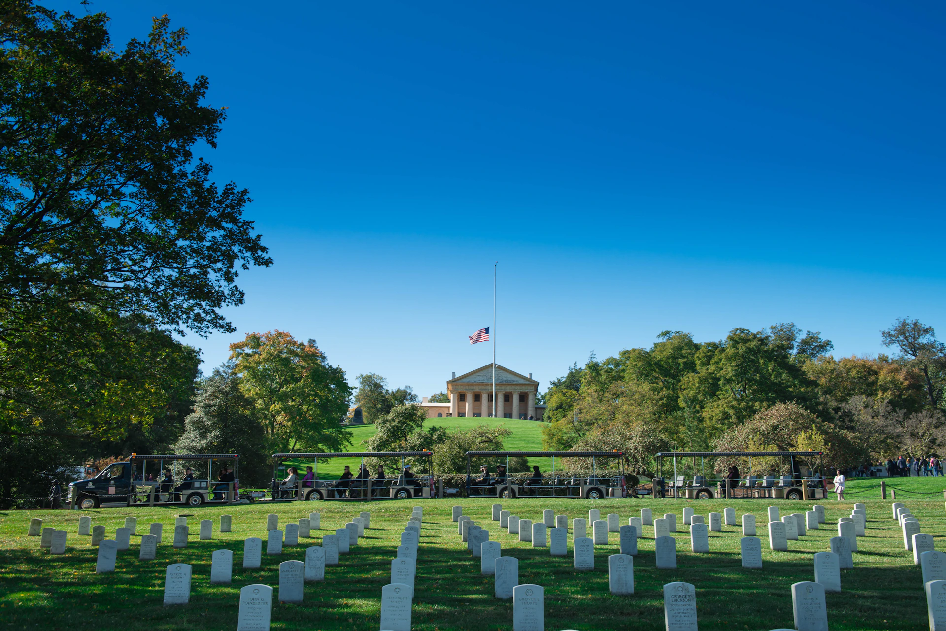 A national cemetery with rows of white headstones, a flag at half-mast, and a columned building on a hill in the background.