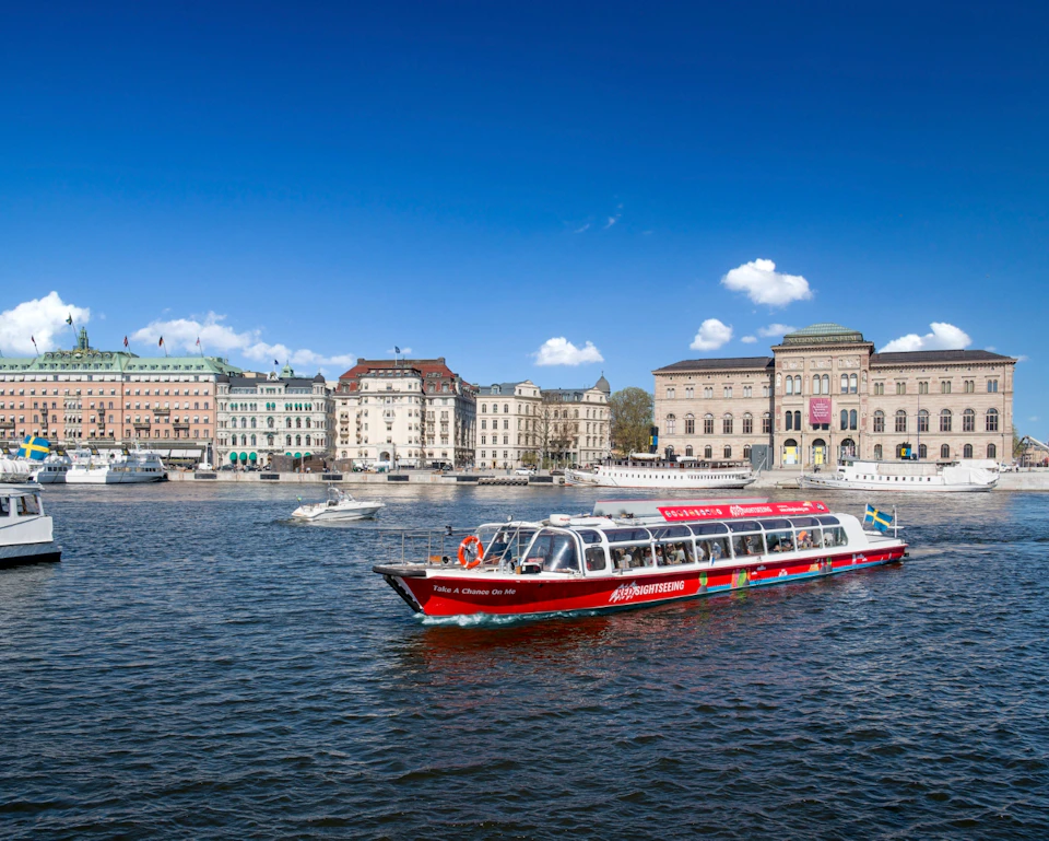 A red sightseeing boat on a river, with historic buildings and a bright blue sky in the background.