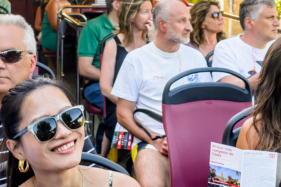 People enjoying a guided tour on an open-top bus in a sunny city, with historic buildings in the background.