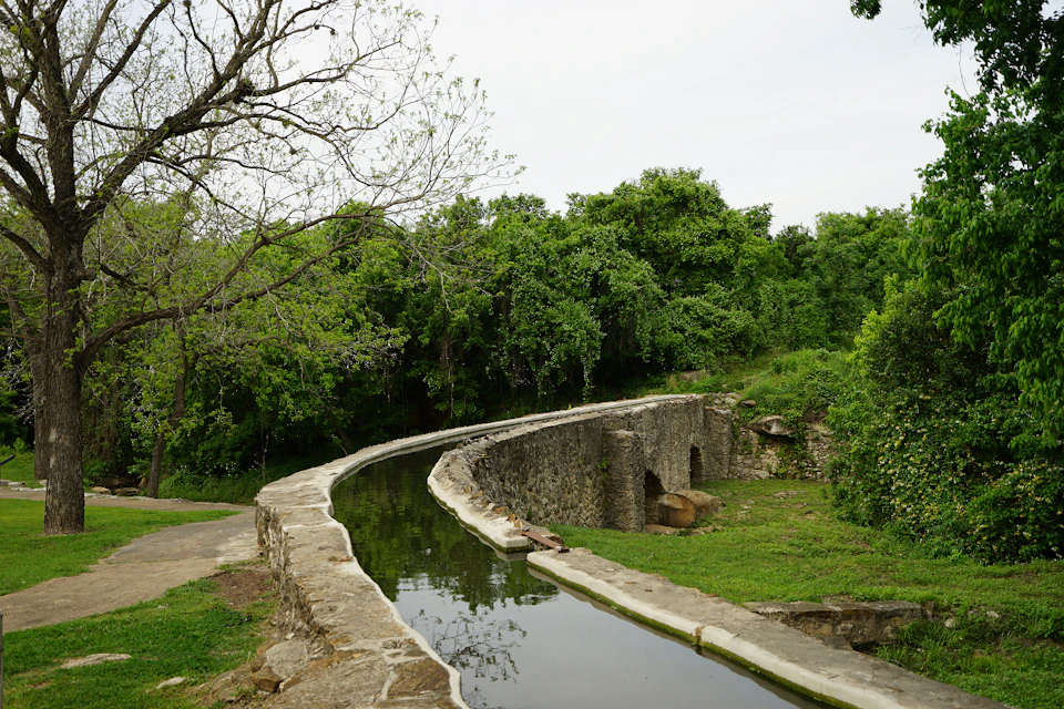 A stone-lined water channel curves through a lush, green forested area with an overcast sky.