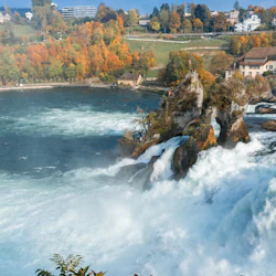 Rusande vattenfall med dimma vid basen, omgiven av höstträd och flera byggnader på kullarna i bakgrunden.