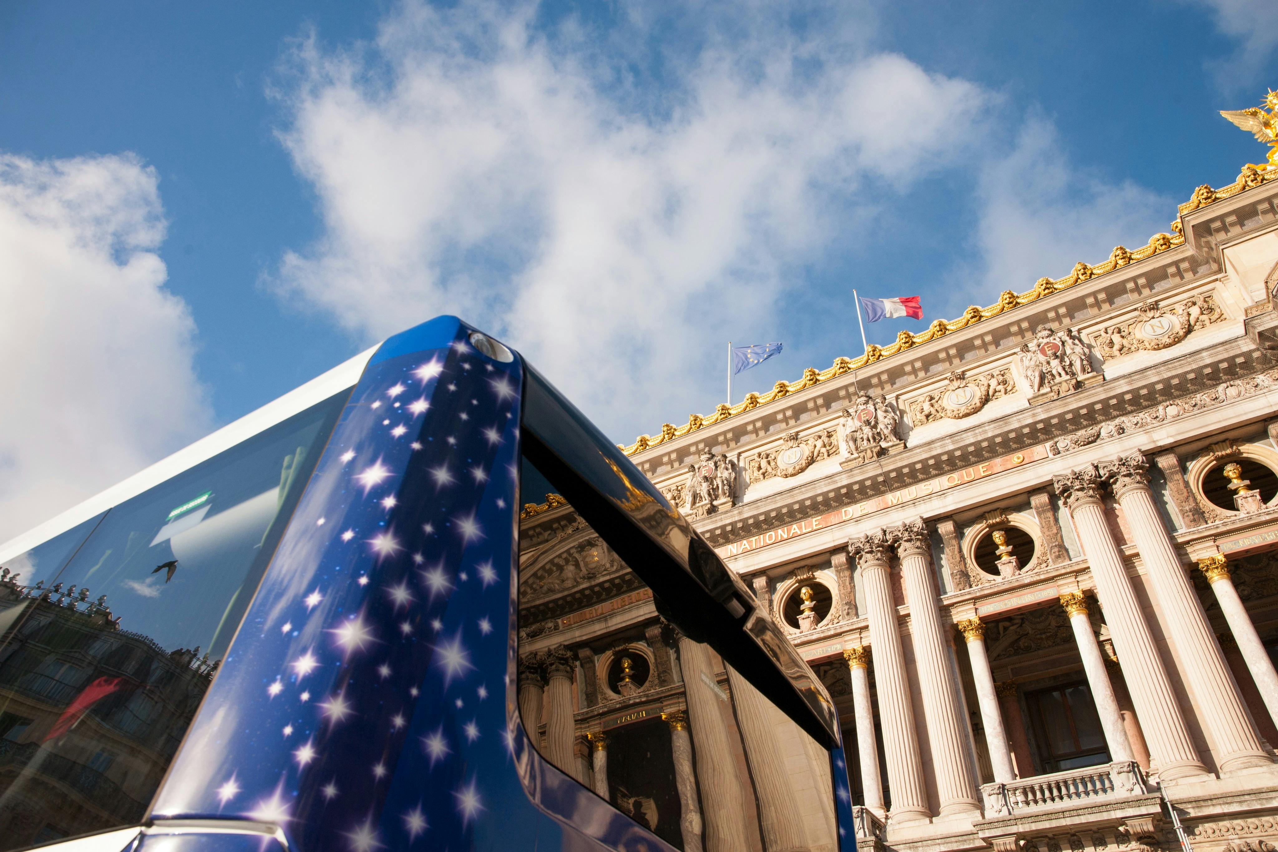 파란 하늘 아래 기둥과 프랑스 국기로 장식된 클래식 건물 앞에 별이 빛나는 디자인의 파란색 버스가 있습니다.
