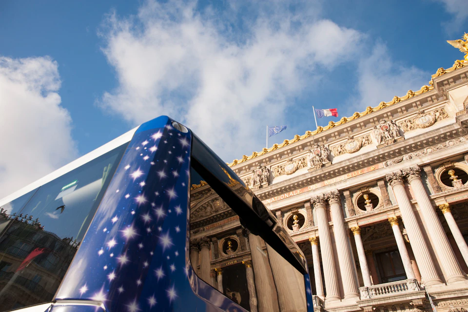 A blue bus with a starry design in front of a classical building adorned with columns and French flags, under a blue sky.