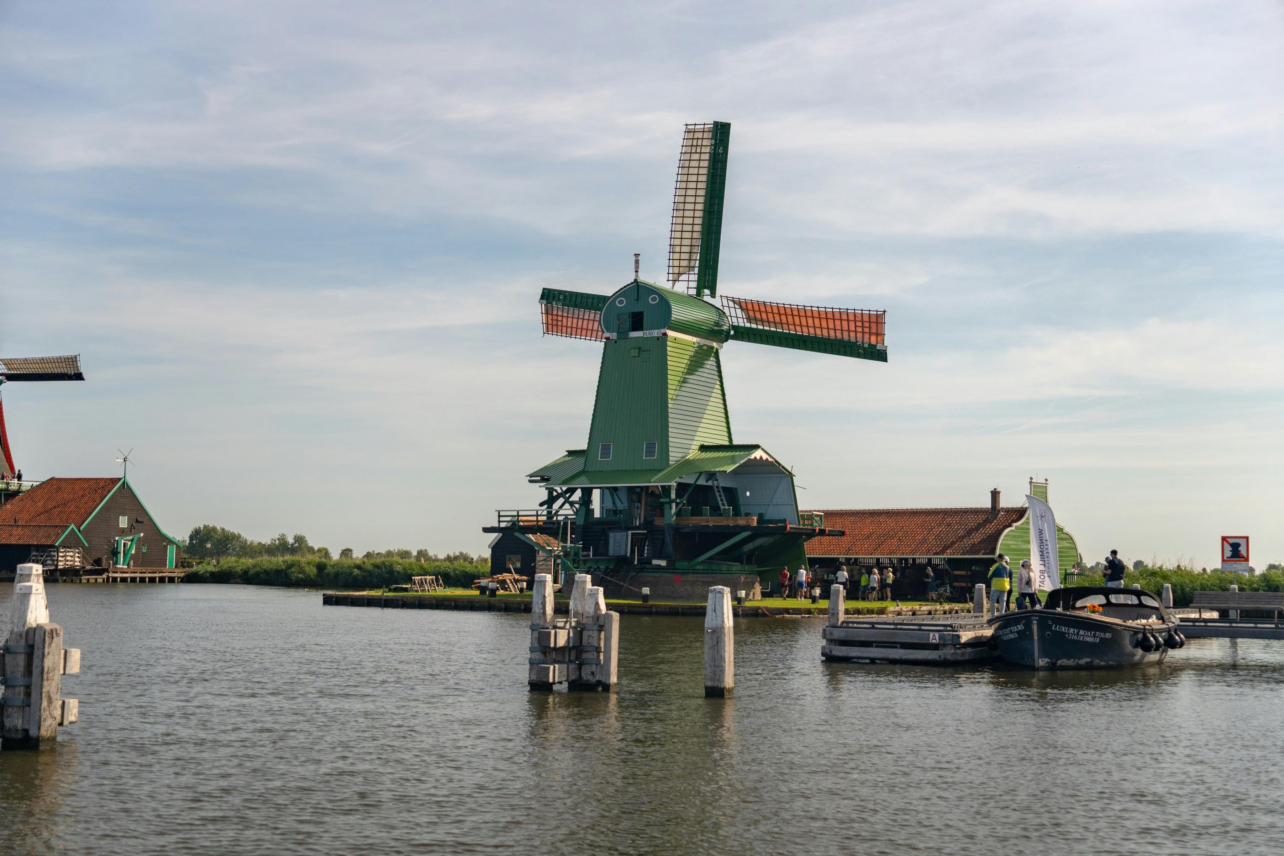 A green windmill by a river with people nearby, boats docked, and a clear sky in the background.