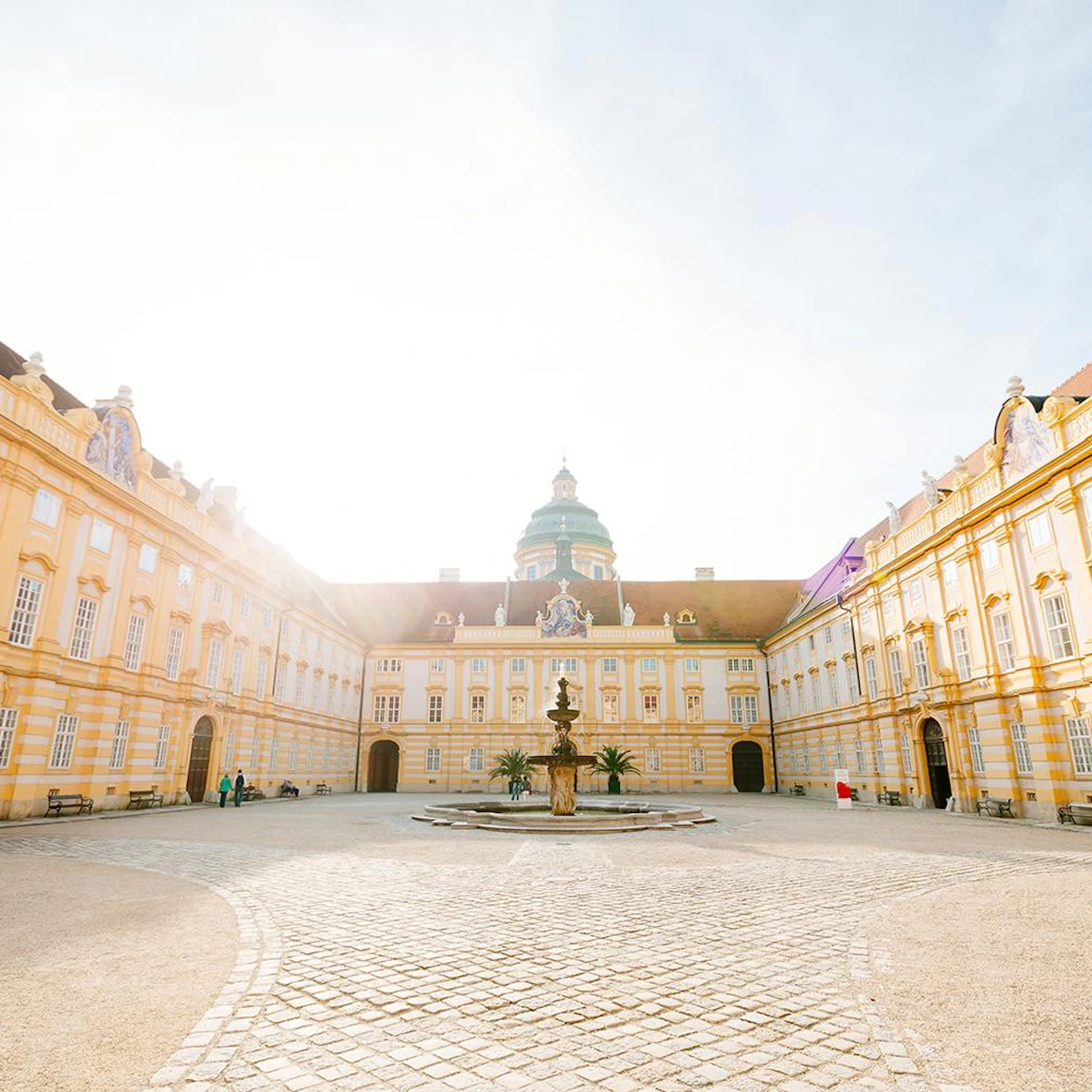 A grand courtyard with a central fountain, surrounded by symmetrical yellow buildings and arched windows, under a bright sky.