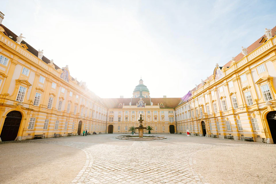 A grand courtyard with a central fountain, surrounded by symmetrical yellow buildings and arched windows, under a bright sky.
