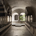 Stone corridor with striped, arched columns, leading to a courtyard with a green tree. Steps descend from the arched opening.