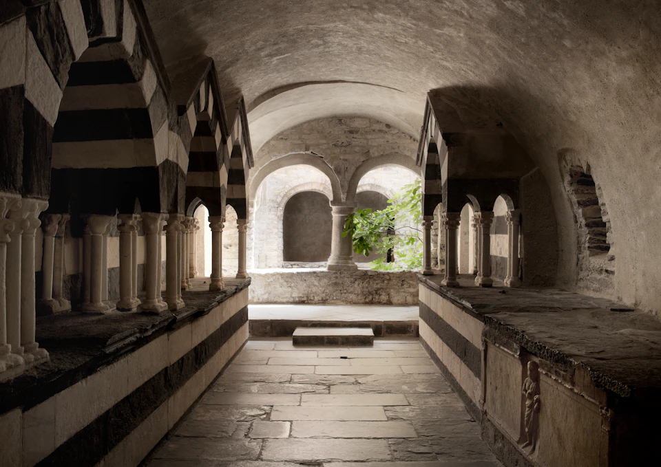 Stone corridor with striped, arched columns, leading to a courtyard with a green tree. Steps descend from the arched opening.