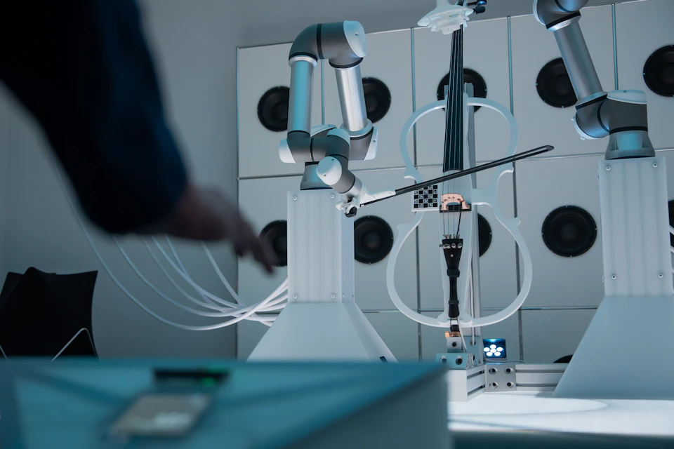 A robotic arm plays a violin in a high-tech setting, with speakers in the background and a person partially visible on the left.