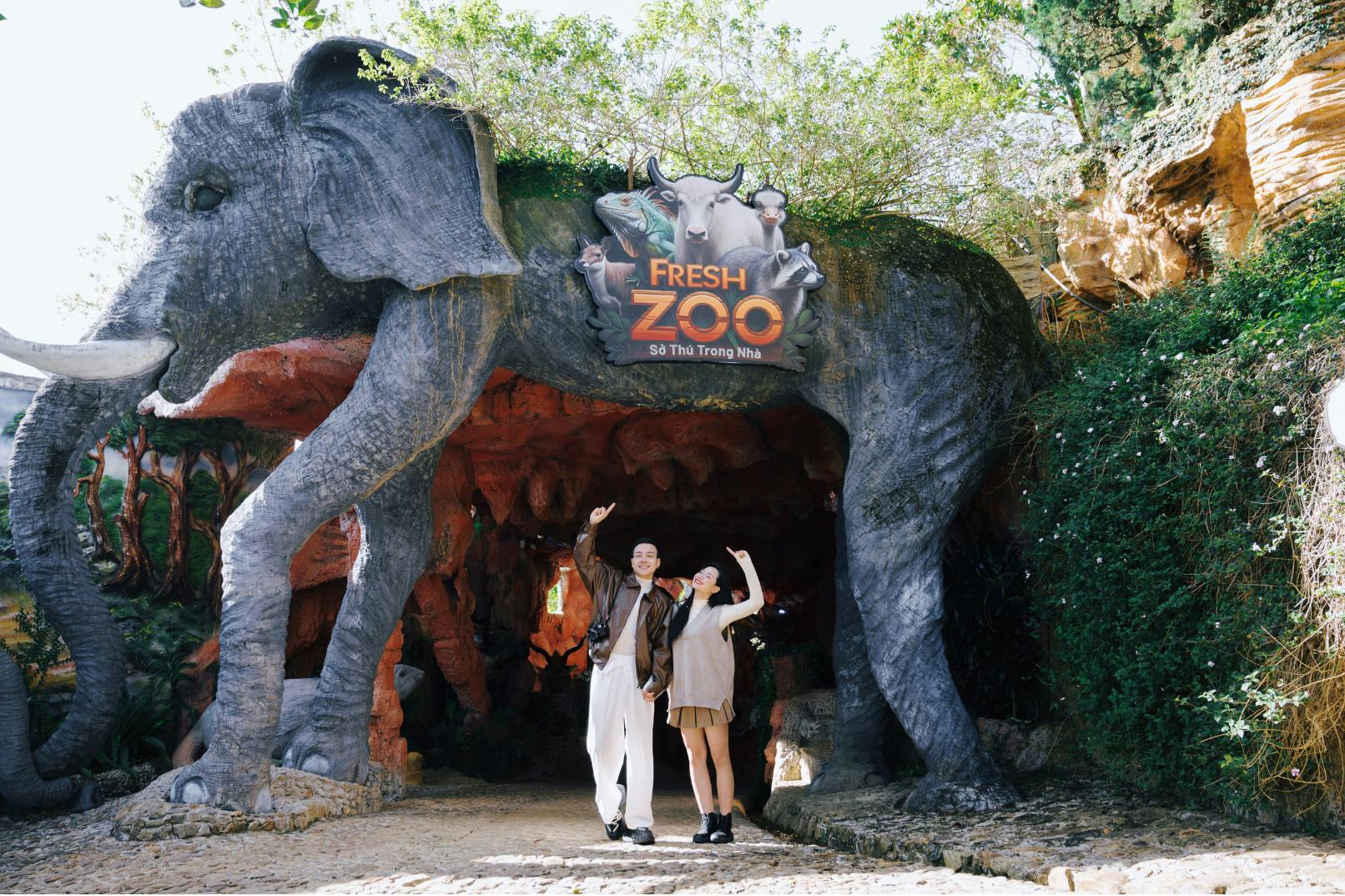 Two people posing at the entrance of a zoo with a large elephant sculpture and a sign reading "Fresh Zoo."