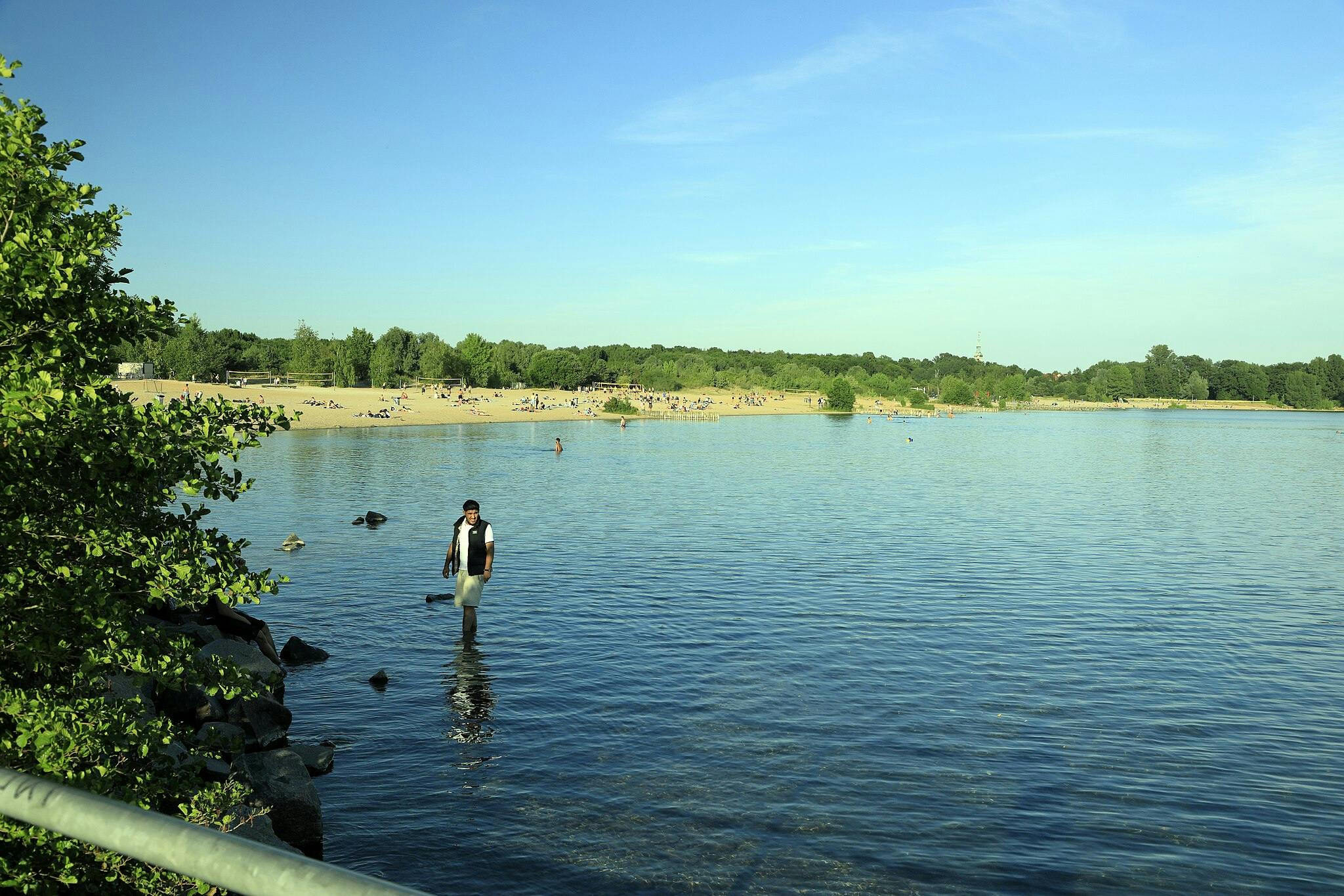 A person wades in a calm lake near a sandy beach with scattered people, adjacent to trees and under a clear blue sky.