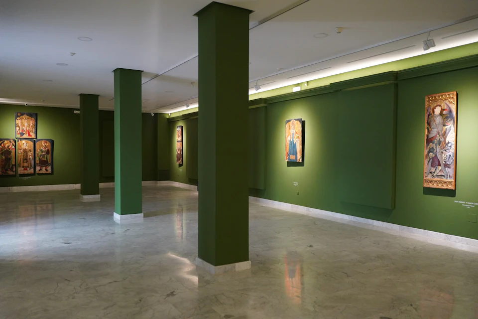An art gallery with green walls, marble flooring, and framed artworks illuminated by lights on display.