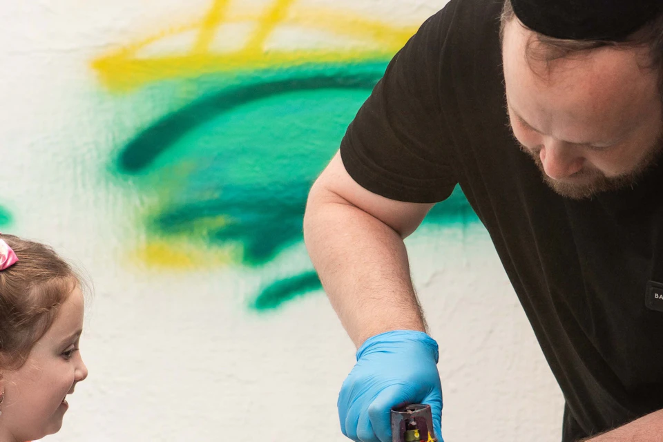 An adult helps a child with spray painting. Both wear blue gloves. Spray cans and colorful graffiti are on the wall behind them.