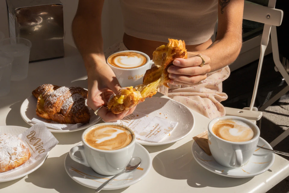 Person tearing a pastry over a plate with two cups of cappuccino on the table.