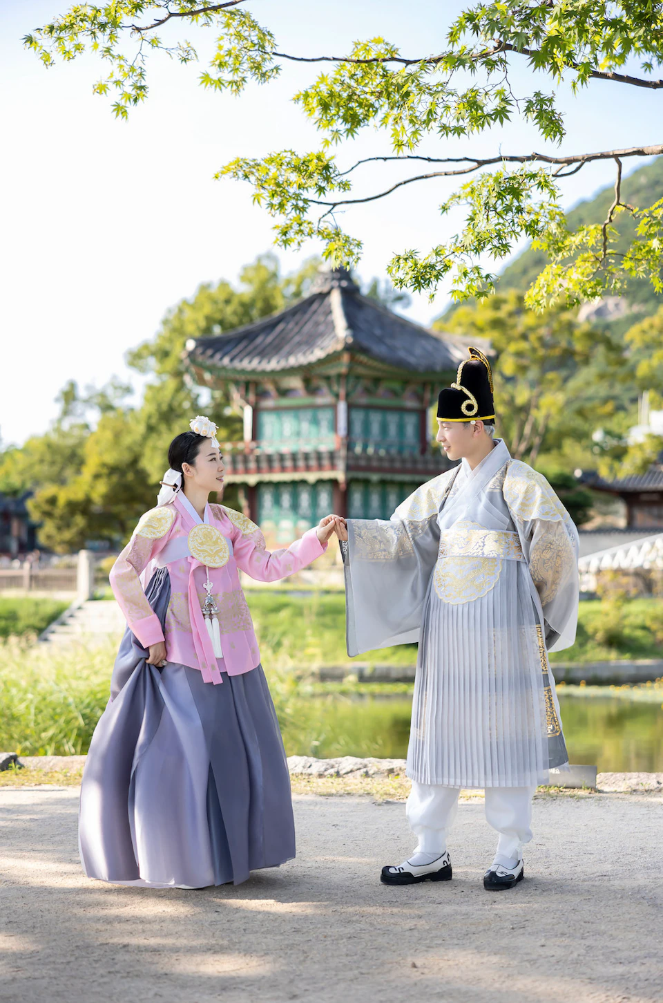 Two people in traditional Korean attire stand outdoors near a pond, with a historical pavilion in the background.