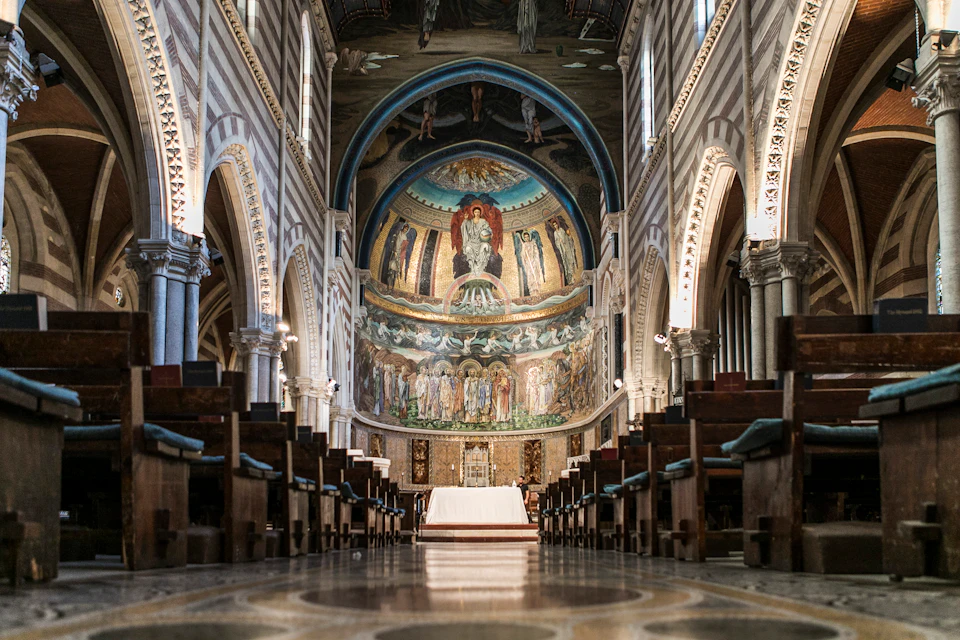 Interior of a grand cathedral with a decorated apse, arched colonnades, pews, and ornate detailing, depicting religious artwork.