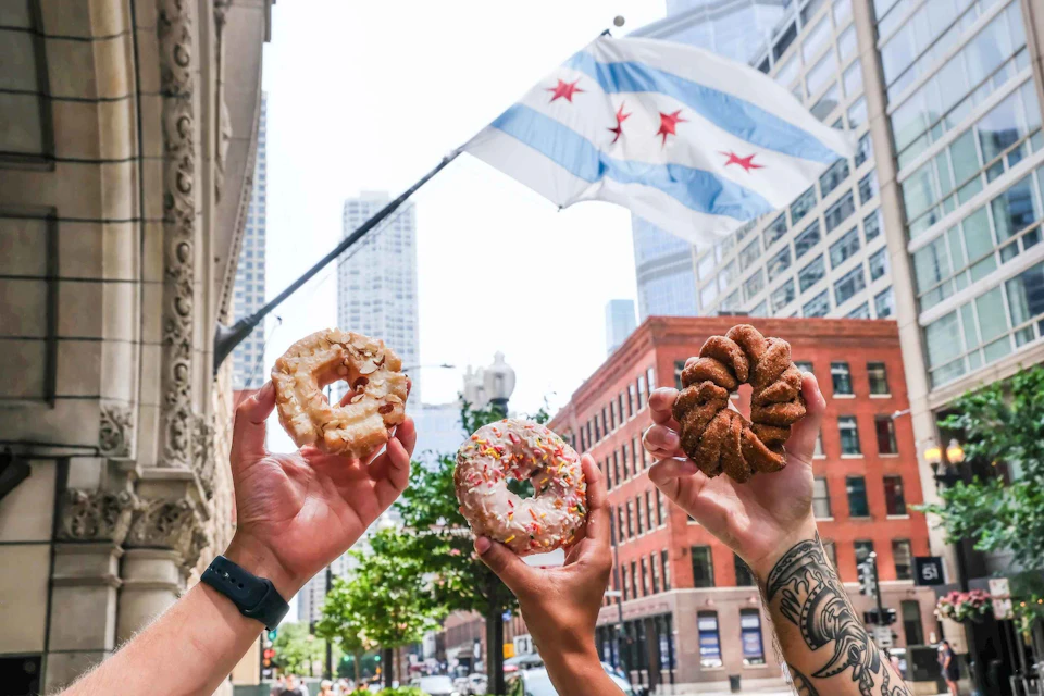 Three hands holding differently decorated donuts with the Chicago flag and city buildings in the background.