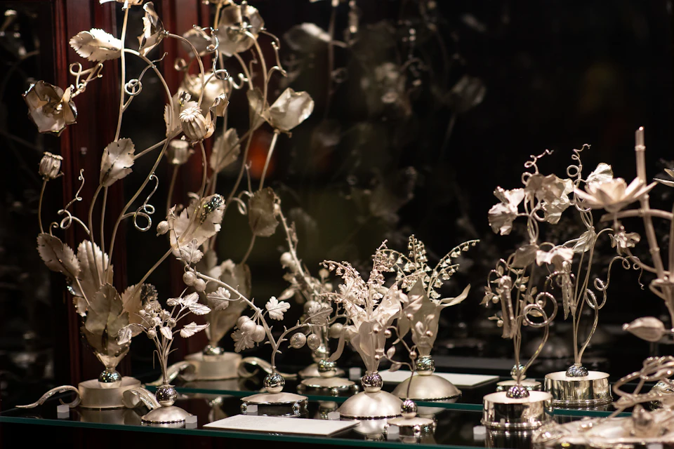 Intricately crafted metallic flower sculptures on a reflective surface, with a dark, blurred background.