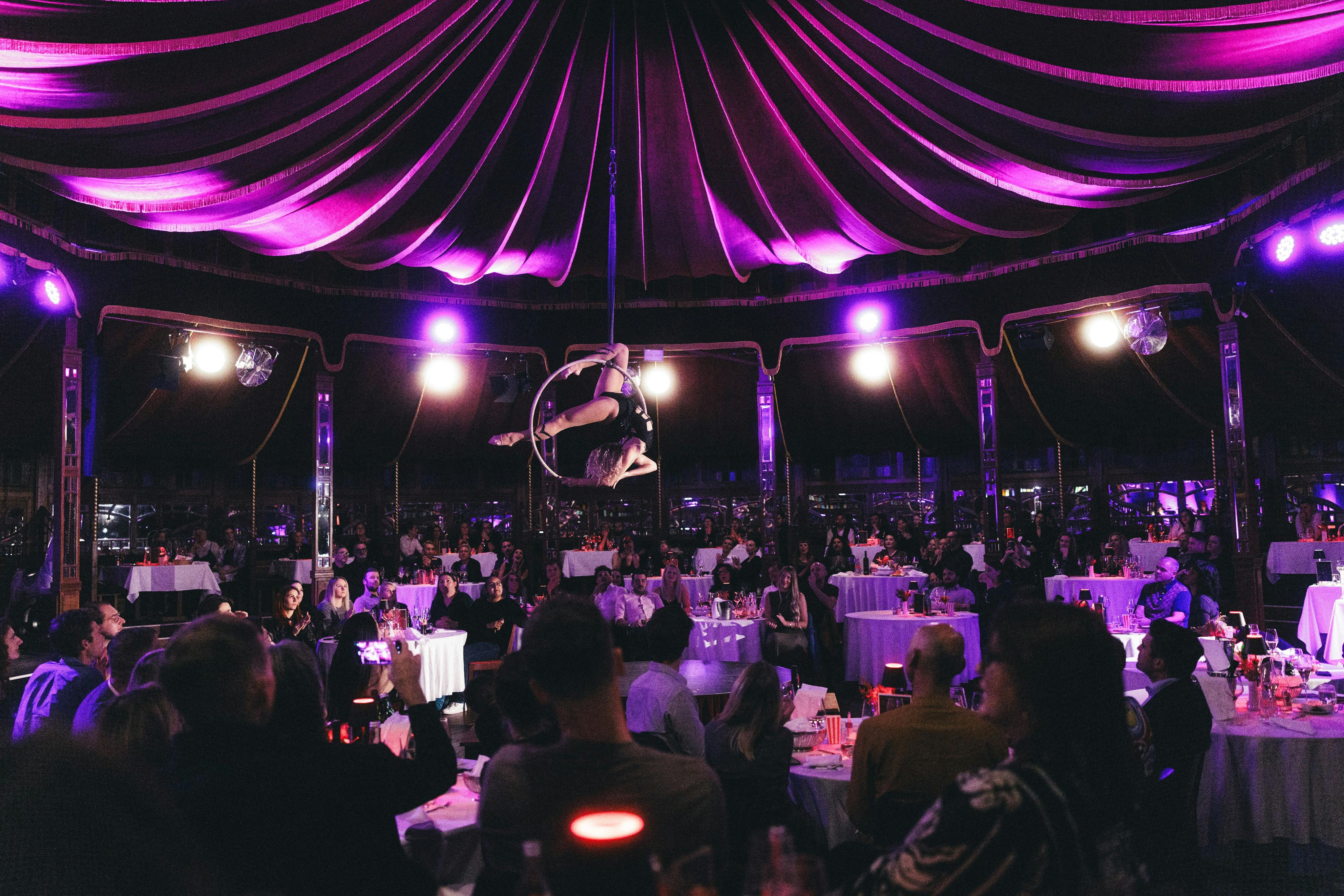 Ο αεροπόρος παίζει σε ένα στεφάνι πάνω από ένα ακροατήριο που κάθεται σε τραπέζια σε έναν αμυδρά φωτισμένο, μοβ χώρο.