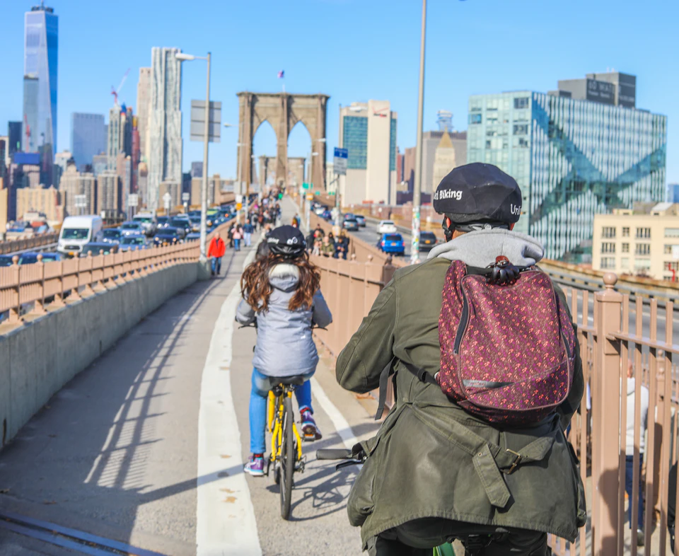People riding bicycles on a path towards the Brooklyn Bridge, with city skyscrapers in the background on a clear day.